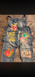 Load image into Gallery viewer, A pair of faded blue denim overalls laid flat on a rustic wooden surface, decorated with colorful embroidered patches including pink roses, yellow text, and floral designs. Sun Faded Trail Overalls by Jaded Gypsy.