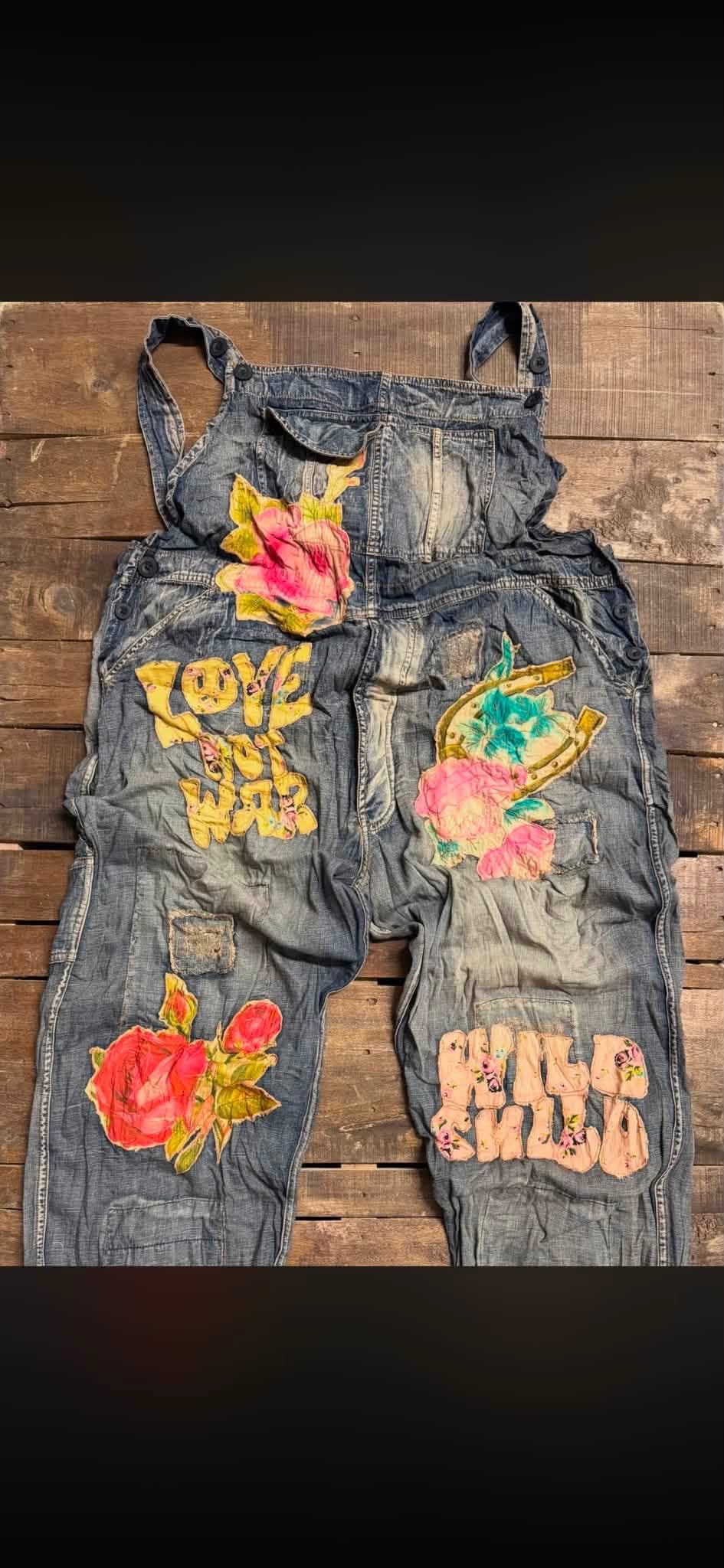 A pair of faded blue denim overalls laid flat on a rustic wooden surface, decorated with colorful embroidered patches including pink roses, yellow text, and floral designs. Sun Faded Trail Overalls by Jaded Gypsy. 